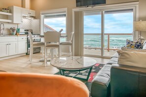 A modern living room with a large window overlooking the sea, a dining area with white chairs, and a kitchen with white cabinets. - Spectacular Ocean Views! Watch Whale Migrations! Dog Friendly! (Depoe Bay)