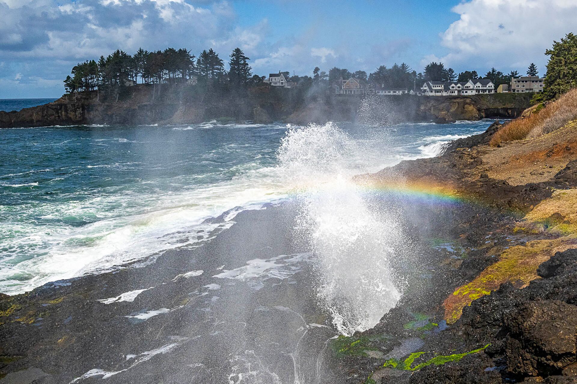 Escena costera con olas rompiendo contra rocas, un arcoíris y casas al fondo.