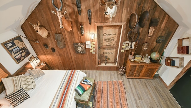 A room with a sloped ceiling, wooden wall decor including animal heads and masks, a bed with patterned bedding, a small wooden cabinet, and a framed picture on the wall. - Suite 4: Outpost · Gorgeous Farm Carriage House Loft (Berryville)