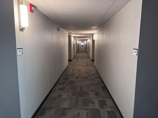 Un long couloir étroit, typique des hôtels, avec un sol recouvert de moquette et des murs sur lesquels sont indiqués les numéros des chambres.