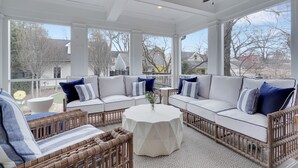 Ein überdachter Patio-Bereich mit einem weißen Ecksofa, Korbmöbeln und einem runden Tisch mit einer Vase.