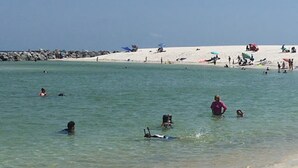 Uma praia com águas cristalinas, pessoas nadando e tomando sol, e uma faixa de areia.