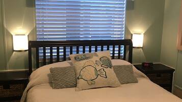 A bedroom with a bed, two bedside lamps, a window with blinds, and a tropical-themed curtain.