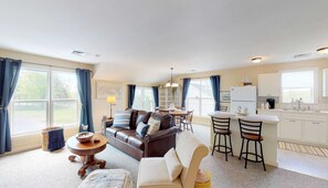 A spacious living room with a brown leather sofa, a white armchair, and a round wooden coffee table. A dining area with a white table and chairs is adjacent to the living room. The kitchen features white cabinets and a double sink. - 1 Bdrm Pet friendly Rental/ Walkable to Village (Skaneateles)