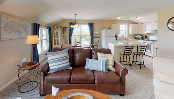 A cozy living room with a brown leather sofa, a dining area with a table and chairs, and a kitchen with white cabinets and appliances.