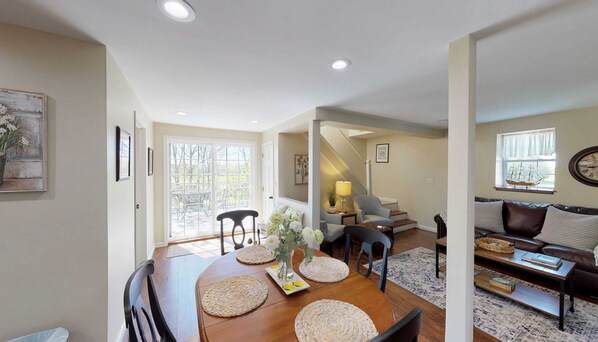 A dining area with a wooden table, woven placemats, and a vase of flowers. There is a staircase leading to a living room with a sofa and a lamp.