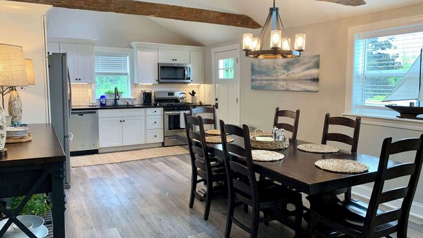 A modern kitchen with white cabinets, a stainless steel refrigerator, and a microwave. There is a dining area with a wooden table and chairs, and a chandelier above. - 2 Bedroom/Pet Friendly/short walk to Village (Skaneateles)