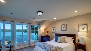 A bedroom with a large bed, a nightstand, a chair, and a view of the water through large windows. - Level Lakefront on Owasco Lake! Sleeps 14! (Auburn)