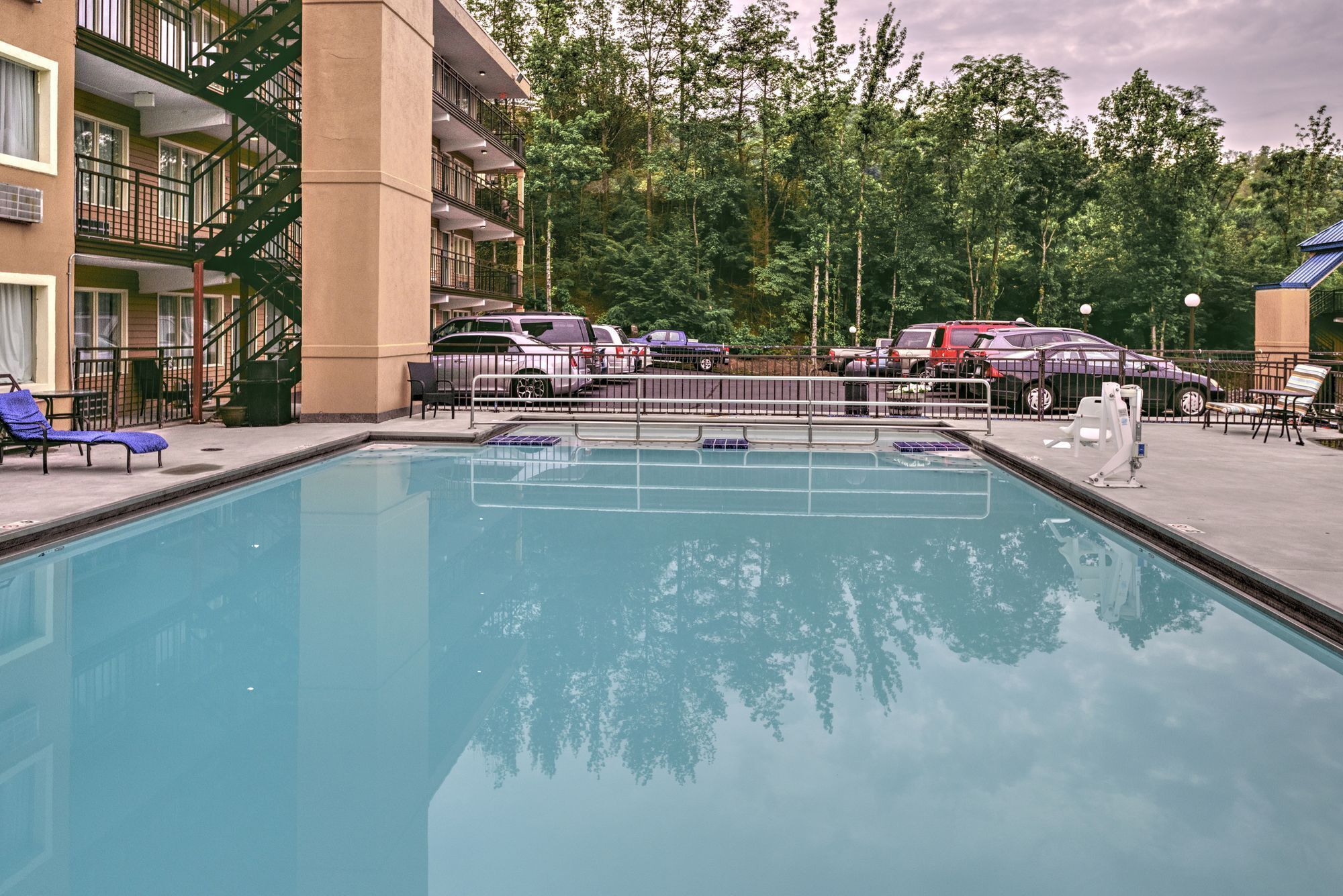 Una piscina con sillas de descanso, un estacionamiento cercado y un edificio de varios pisos con balcones.