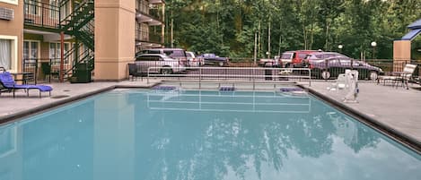 Una piscina con sillas de descanso, un estacionamiento cercado y un edificio de varios pisos con balcones.