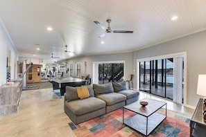 A spacious living area with a gray sofa, a coffee table, and a large window offering a view of a body of water. - Lake Front Home with Pool, Dock, Views, Sports Court, & Beautiful Sunsets! (Lago Vista)