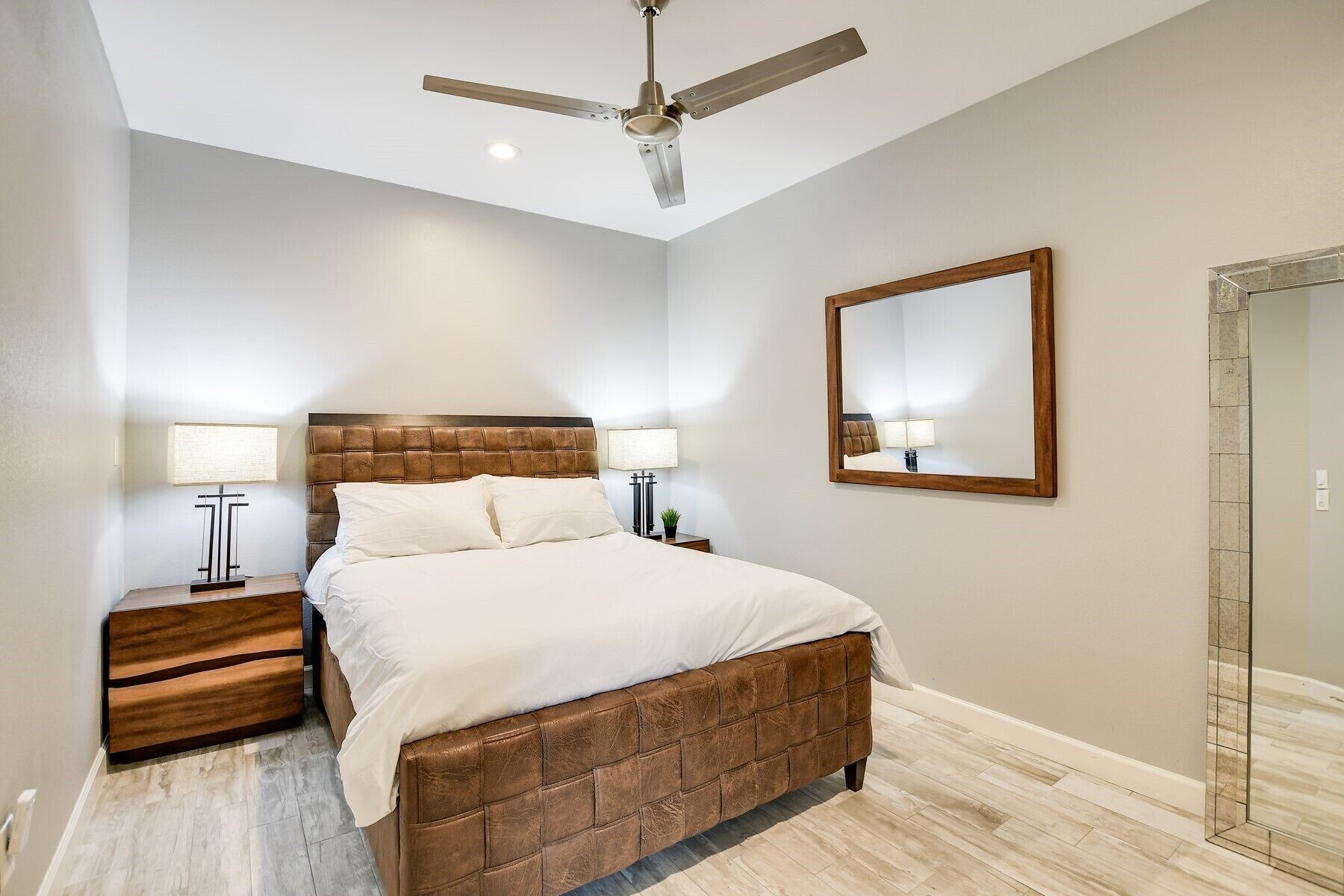 A bedroom with a wooden headboard bed, two bedside tables with lamps, a ceiling fan, and a large mirror on the wall.
