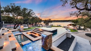 A pool area with a waterfall feature, surrounded by stone steps and benches, with a view of a lake and trees at sunset. - Lake Front Home with Pool, Dock, Views, Sports Court, & Beautiful Sunsets! (Lago Vista)