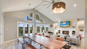 A spacious living area with a large wooden dining table, comfortable seating, a fireplace, and a television mounted above it. - Lake Front Home with Pool, Dock, Views, Sports Court, & Beautiful Sunsets! (Lago Vista)