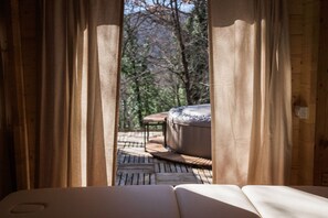 Una terraza de madera con un sofá y una mesa, cortinas enmarcan vistas a árboles.
