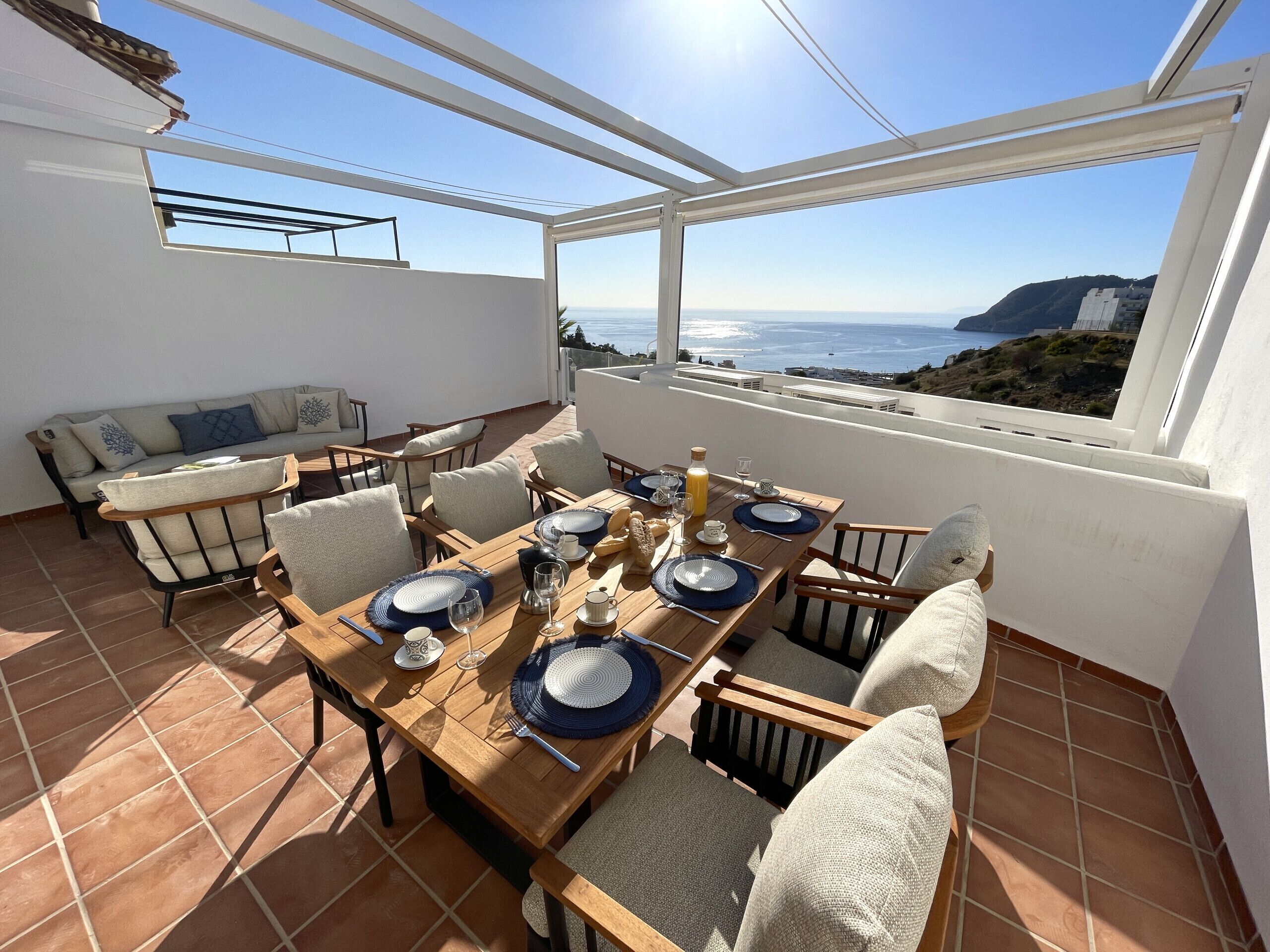 Un patio soleado con una mesa de madera preparada para comer, un sofá y vistas al mar.