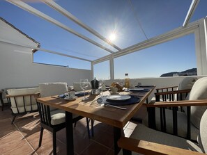 A sunlit outdoor dining area with a wooden table set for two, surrounded by cushioned chairs, and overlooking a scenic landscape. - Stunning 3 bedroom house with communal pool and views (La Herradura)