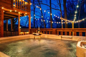 A hot tub area with string lights, wooden deck, and a view of trees.