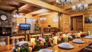 A cozy log cabin interior with a fireplace, wooden walls, a dining table set for a meal, and a Christmas tree. - The Hilltop Chalet : Location, Views, Hot tub, Fire Pit, Game Room! (Gatlinburg)