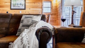 A cozy cabin interior with a leather sofa, a white knitted throw, a framed picture on the wall, and a lamp with a bird decoration. - The Hilltop Chalet : Location, Views, Hot tub, Fire Pit, Game Room! (Gatlinburg)