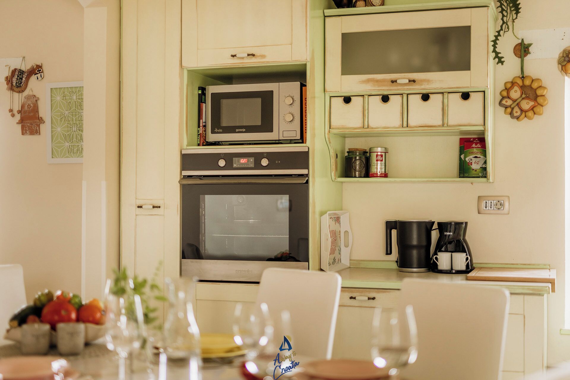 Een keuken met een magnetron, oven en koffiezetapparaat. Er zijn kasten, een fruitschaal en een tafel met stoelen.