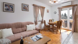 Ein Wohnzimmer mit einer Couch, einem Esstisch mit Stühlen, einem Couchtisch mit einer Weinflasche und Gläsern sowie Blick ins Freie durch eine Glastür.
