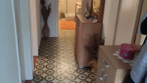 Un couloir étroit avec des carreaux de sol à motifs, une armoire en bois dotée de tiroirs, un miroir et une petite table sur laquelle est posé un objet décoratif.