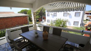 Une terrasse extérieure avec une table et des chaises, un pot de fleurs et une vue sur un bâtiment blanc doté de fenêtres et d’un balcon.