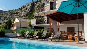 Ein Poolbereich mit Liegestühlen und einem Tisch, ein Gebäude mit einem Balkon und eine Bergkulisse.