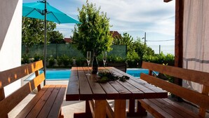 Ein Holztisch mit Bänken, einer Flasche und zwei Gläsern, draußen aufgestellt, mit Schwimmbad und Grünpflanzen im Hintergrund.