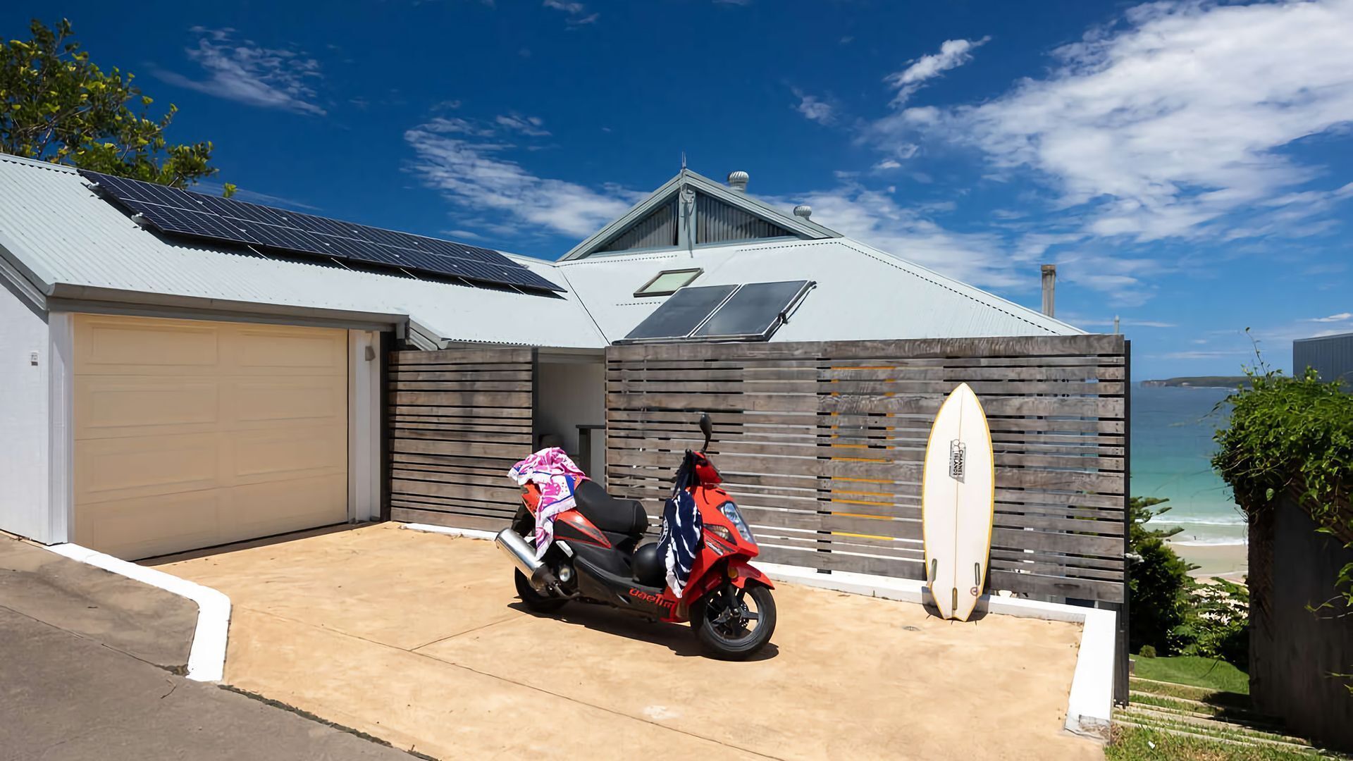 A modern house with a garage, a red scooter, and a surfboard leaning against a wooden fence.
