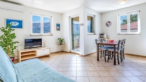 A bright living room with a TV, a sofa, a dining table with chairs, and large windows offering views of the outdoors. - Nikolina B1 - Charming 2-Bedroom Haven for Four, Pet-Friendly with Parking (Baška)