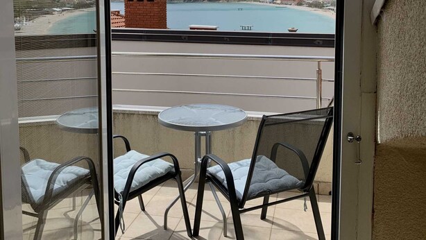A balcony with a glass door, offering a view of a beach, mountains, and a small brick structure. - Nikolina Bon Bon Happy Stay Center – Studio with Terrace & Sea view (Baška)