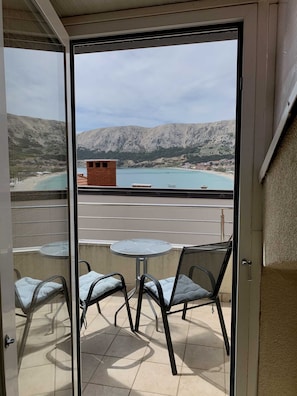 A balcony with a glass door, offering a view of a beach, mountains, and a small brick structure. - Nikolina Bon Bon Happy Stay Center – Studio with Terrace & Sea view (Baška)