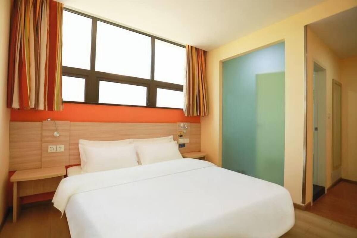 A hotel room with a large bed, a wooden headboard, a nightstand, a window with curtains, and a door to another room.