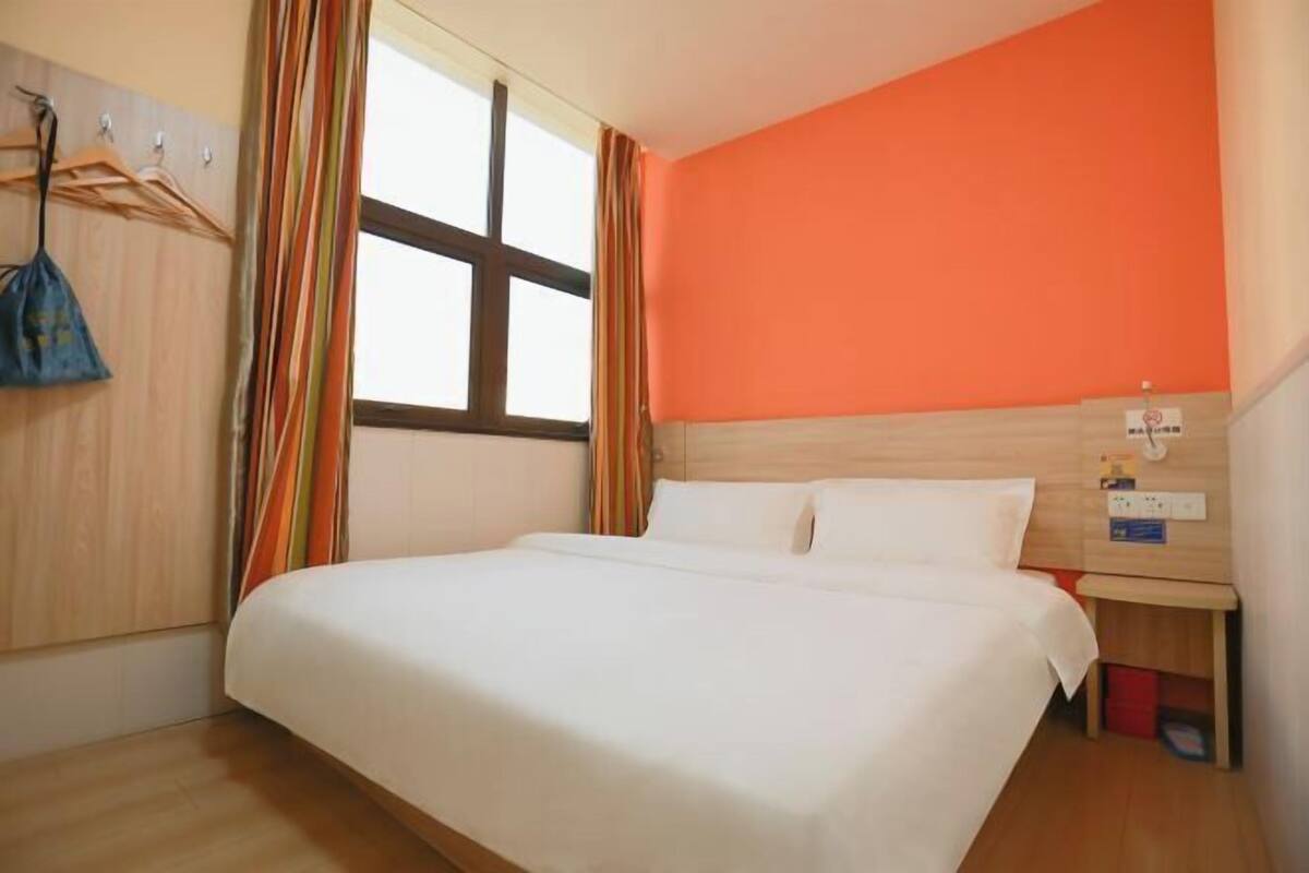 A hotel room with a bed, wooden headboard, a window with curtains, a wall clock, and a small bedside table.