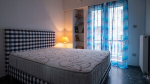 Ein Schlafzimmer mit kariertem Kopfteil, einem Bett mit gemustertem Überwurf, einer Nachttischlampe, einem Regal mit Büchern und Bildern und einem Fenster mit blauen Vorhängen.