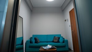 Ein smaragdgrünes Sofa mit gemusterten Kissen, ein Couchtisch aus Holz mit einer Zeitschrift und eine Deckenleuchte in einem Raum mit weißen Wänden und einer Holztür.