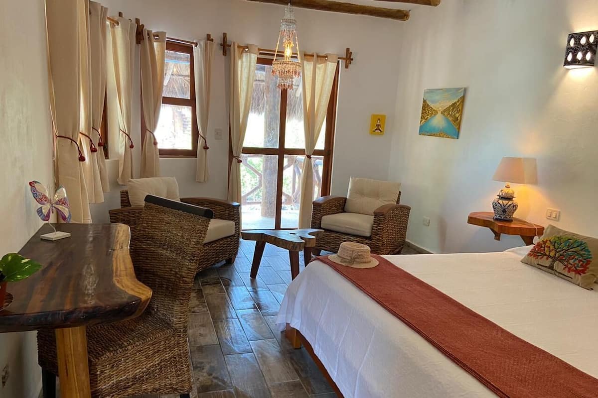 Ein gemütliches Zimmer mit einem Bett, zwei Korbstühlen, einem kleinen Tisch und einer Kandelaber.