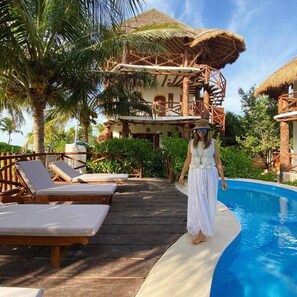 Ein Resort mit Holzterrasse, Liegestühlen, einem Pool und einem Gebäude mit Reetdach.