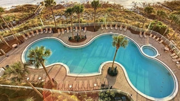 Una piscina de forma peculiar rodeada de sillas de descanso y palmeras, con vista a una playa.