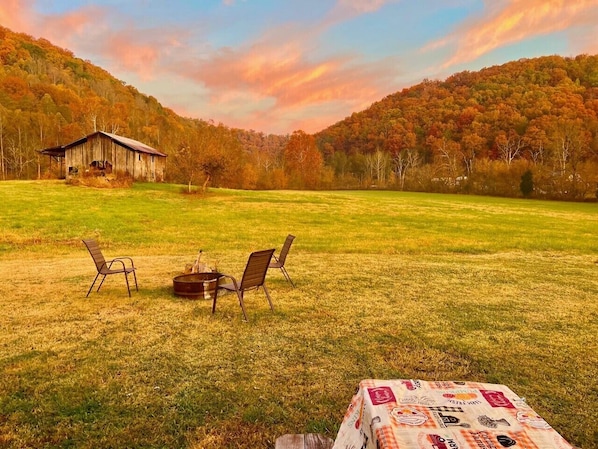 A rustic outdoor setting with a wooden table and chairs, a small wooden structure, and a grassy field with trees in the background.