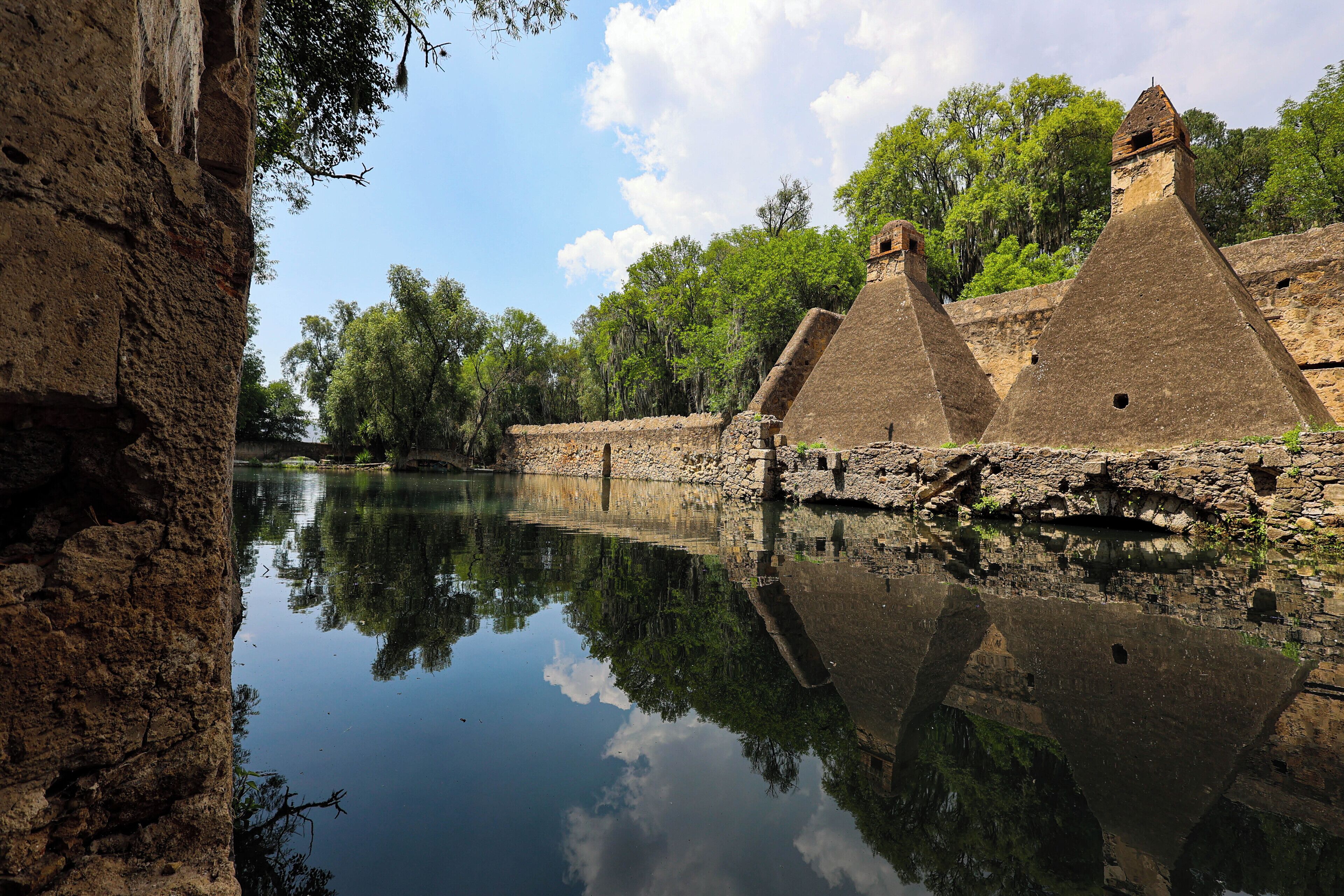 Une structure en pierre qui se reflète dans l’eau, entourée d’arbres.