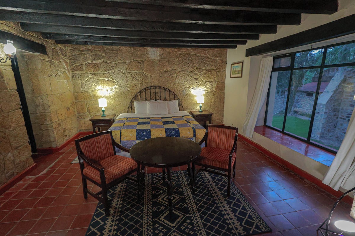 Ein Schlafzimmer mit Steinwand, einem Bett mit kariertem Überwurf, zwei Nachttischlampen, einem runden Tisch und zwei Stühlen.