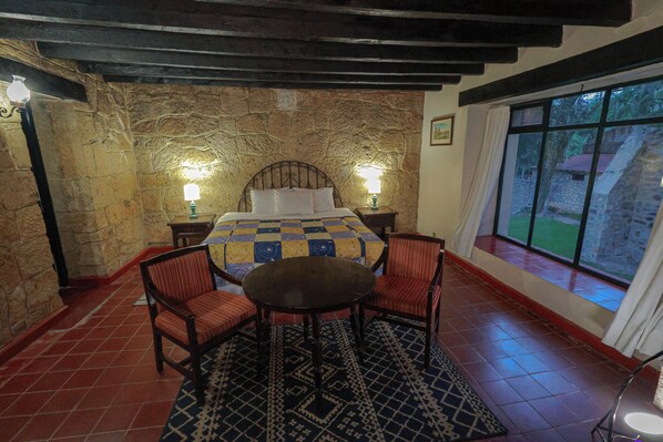 Un dormitorio con pared de piedra, una cama con colcha a cuadros, dos lámparas de noche, una mesa redonda y dos sillas.