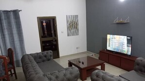Een woonkamer met een grijze zithoek, een salontafel, een televisie op een standaard en een deur met een ruit.