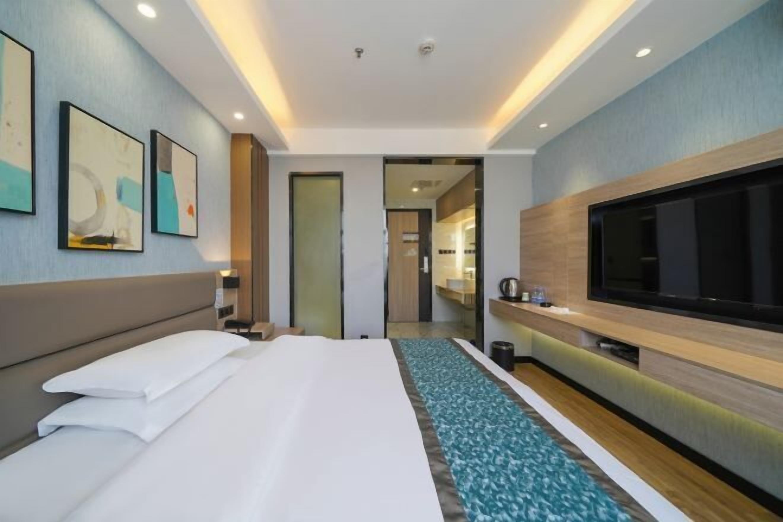Ein modernes Hotelzimmer mit einem großen Bett, einem Flachbildfernseher, einem kleinen Schreibtisch und einem durch eine Glastür einsehbarer Badezimmer.