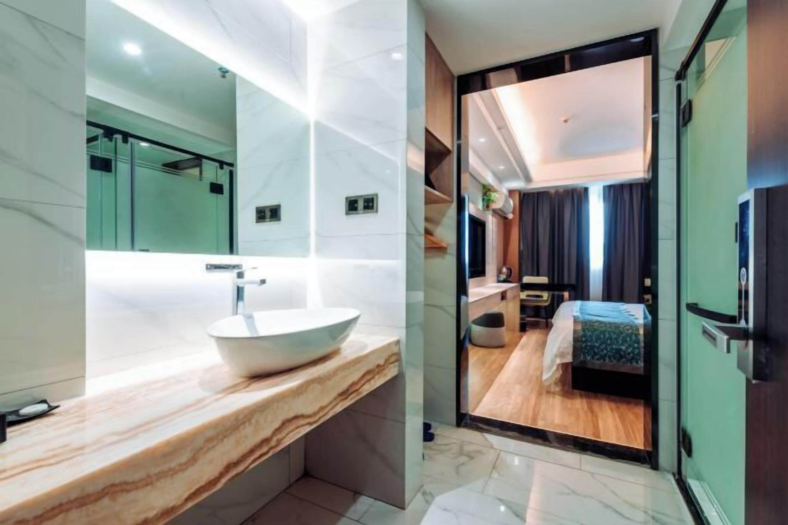 Un baño moderno con tocador de mármol, un espejo grande y vistas a un dormitorio con cama y una mesita.