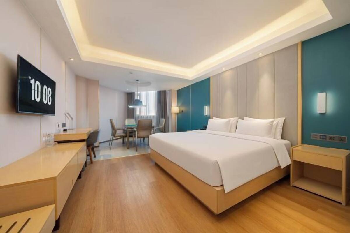 A modern hotel room with a large bed, bedside tables, a desk, and a TV displaying the number 10.