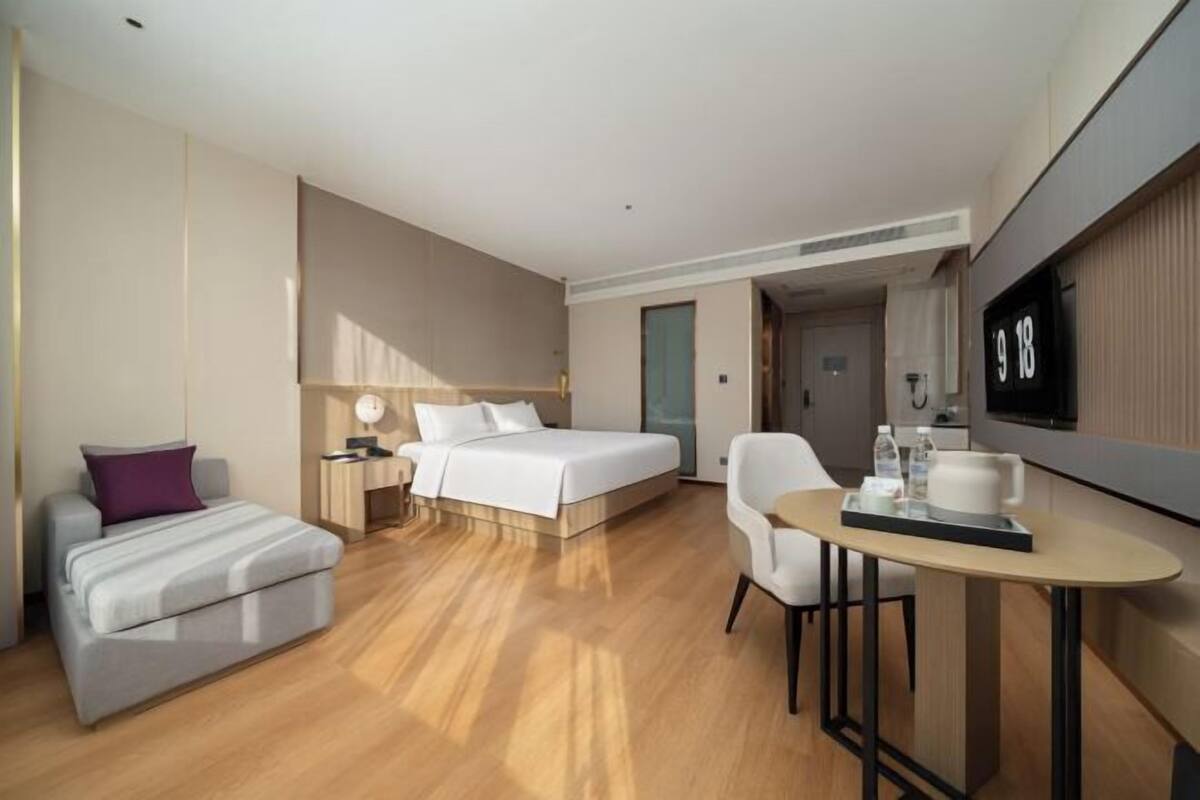 A modern hotel room with a bed, a sofa, a desk with a chair, and a TV on the wall.